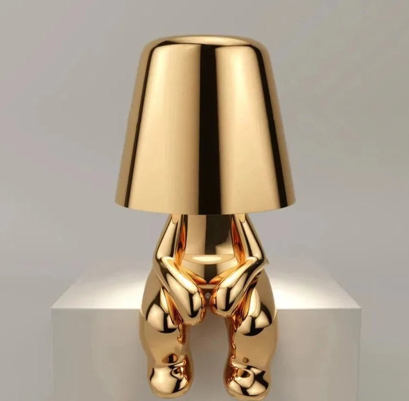 Gold metal table lamp with a cone shaped shade and a base made of sculpted hands resting on a white surface