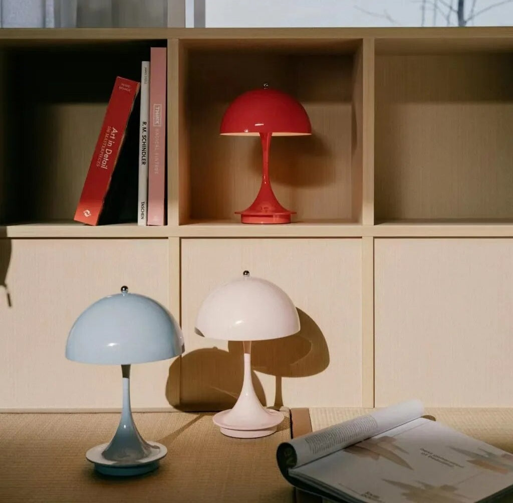 Red LED table lamp with a dome shade and slender stem sits in a wooden cubby unit with blue and pale pink lamps on the surface in front and an open magazine nearby.