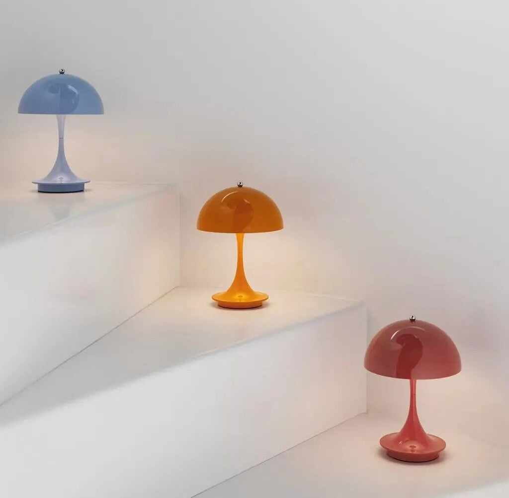 Three colorful dome shaped LED table lamps in blue, orange, and pink with slender curved stems and round bases on white stepped surfaces.