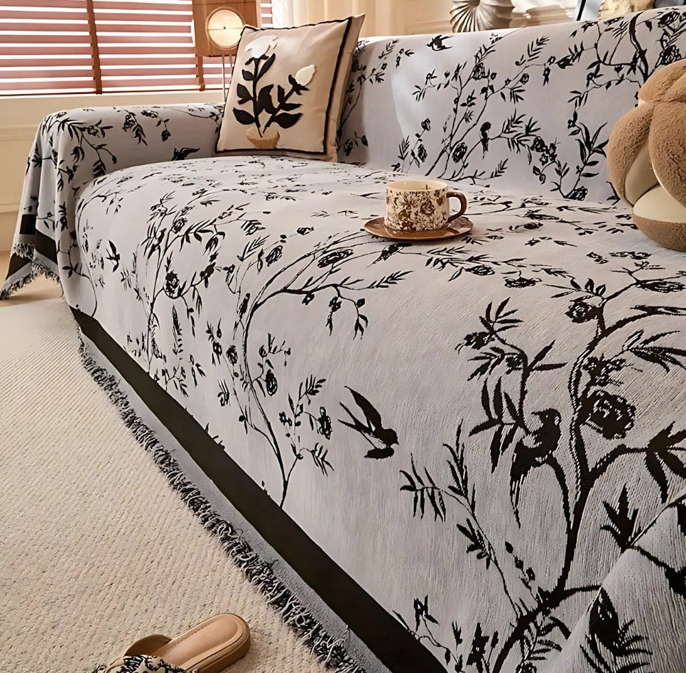 Light gray sofa covered in a chenille jacquard floral throw with tassel trim, a botanical pillow, a coffee cup on a saucer, a teddy bear, and slippers on the light carpet.