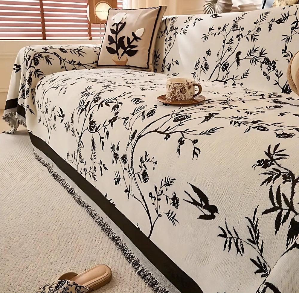 Briarhaven Chenille sofa cover with a black floral jacquard pattern and tassel trim on a cream sofa with a matching pillow and a teacup on a saucer