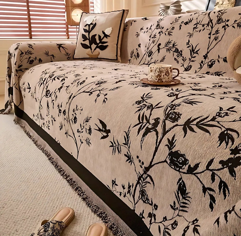 Beige chenille sofa cover with black floral jacquard pattern and tassel trim on a sofa, with a botanical pillow, a teacup on a saucer resting on the cover, and a beige rug and slippers nearby