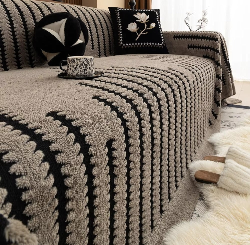 Beige and black tufted knit sofa cover with a linear pattern draped over a sofa, a round black and white pillow and a square black cushion with white floral embroidery, and a teacup on a saucer resting on the cover