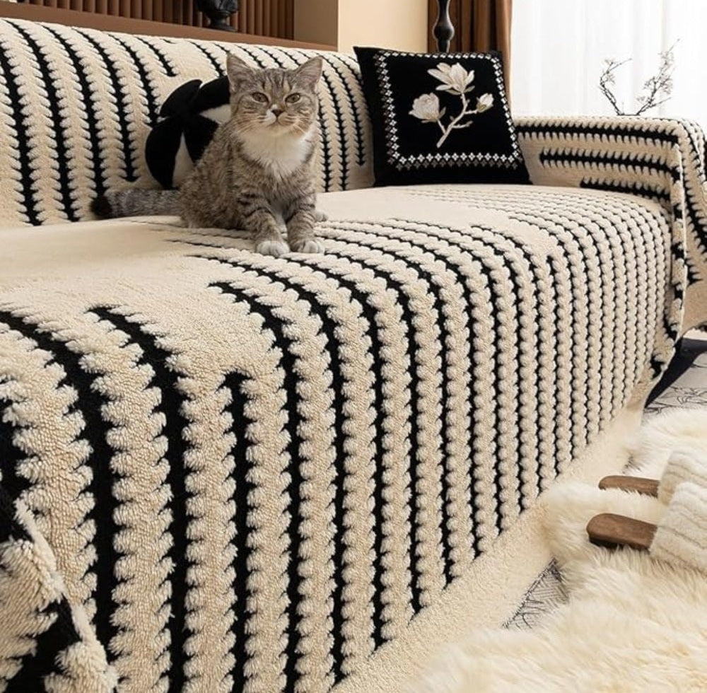 A cat sits on a sofa covered with a plush beige and black tufted striped cover.