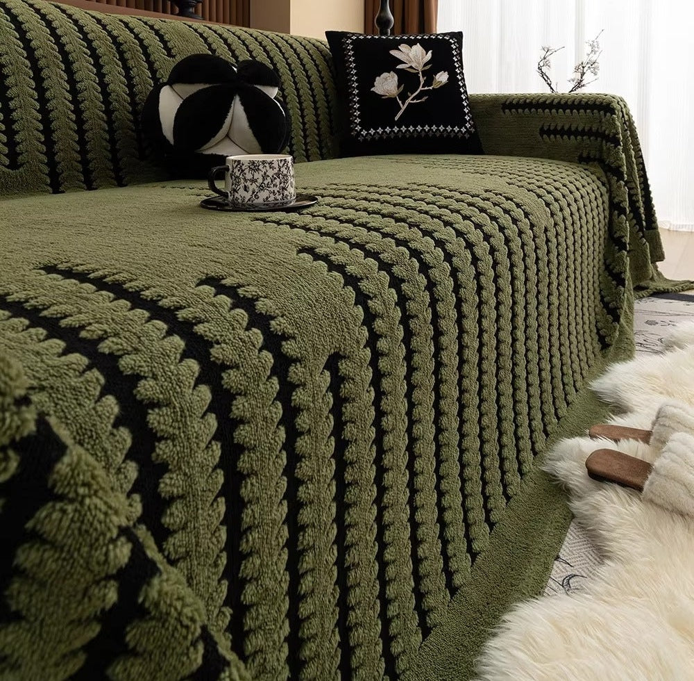Olive green chunky knit sofa cover draped over a sofa with black and white cushions including a square pillow with a white floral design and a round black and white cushion, a cup on a saucer rests on the cover, and a fluffy rug nearby