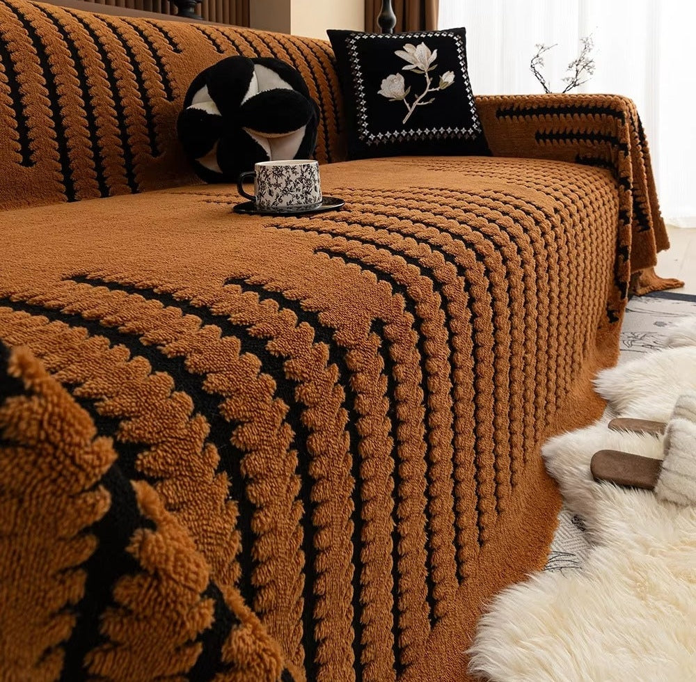 Brown plush knitted sofa cover with a tufted linear pattern draped over a couch, with two decorative cushions one black with white floral embroidery and a round black and white pillow, and a cup on a saucer resting on the cover