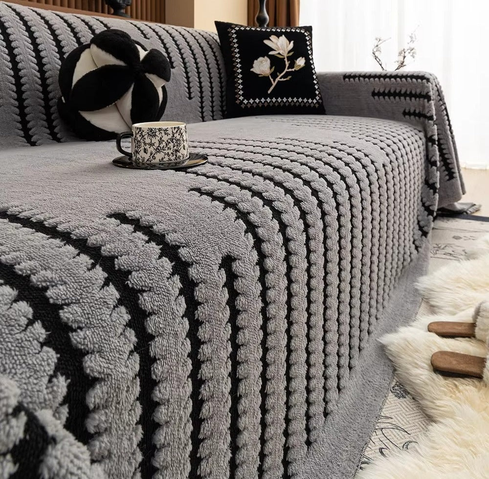 Grey plush sofa cover with a chunky knit pattern on a sofa, a floral embroidered black square pillow and a round black and white pillow on the back, and a teacup on a saucer resting on the cover