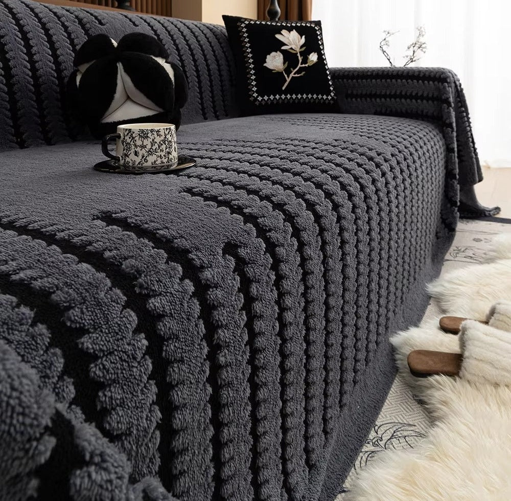Charcoal gray plush sofa cover with a chunky tufted linear knit draped over a sofa, a floral teacup on a saucer rests on the cover, decorative pillows including one with white floral embroidery, and a white fluffy rug with wooden slippers nearby.
