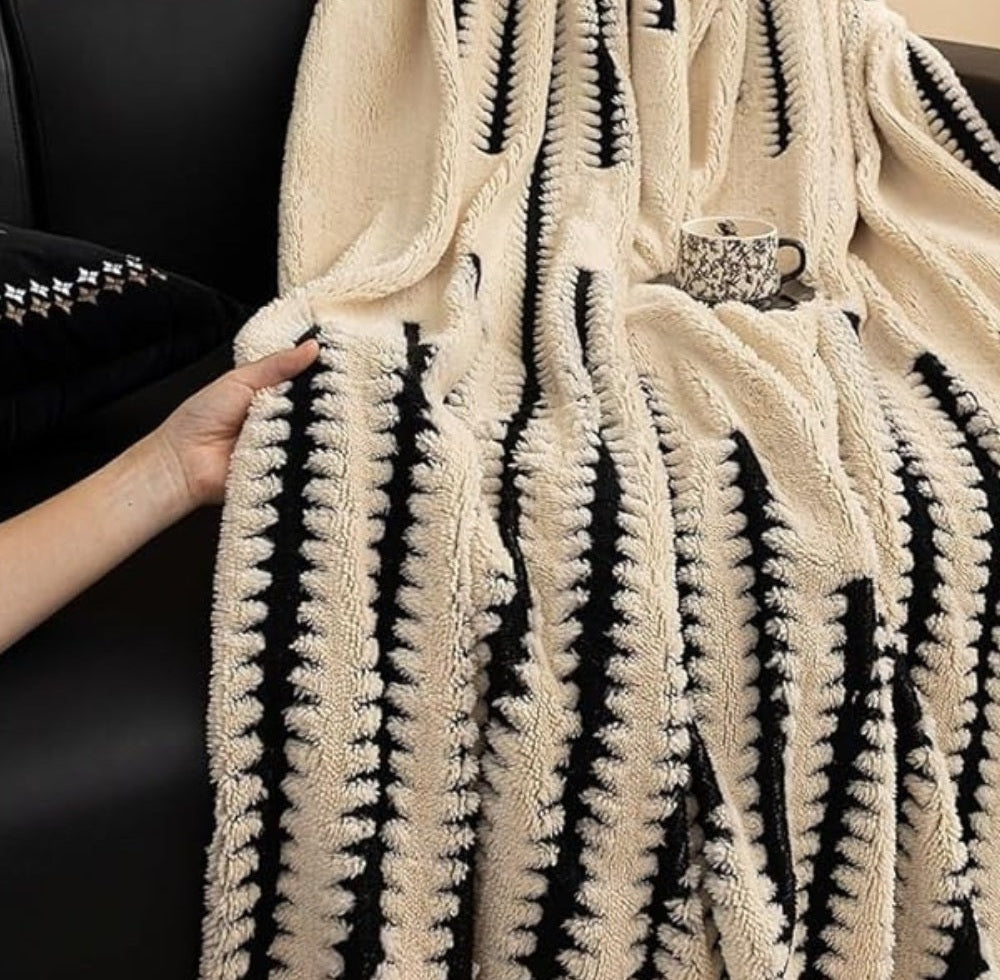 Beige plush throw blanket with tufted black stripes and pom pom fringe draped over a black sofa, a hand holding the edge and a patterned cup sitting nearby
