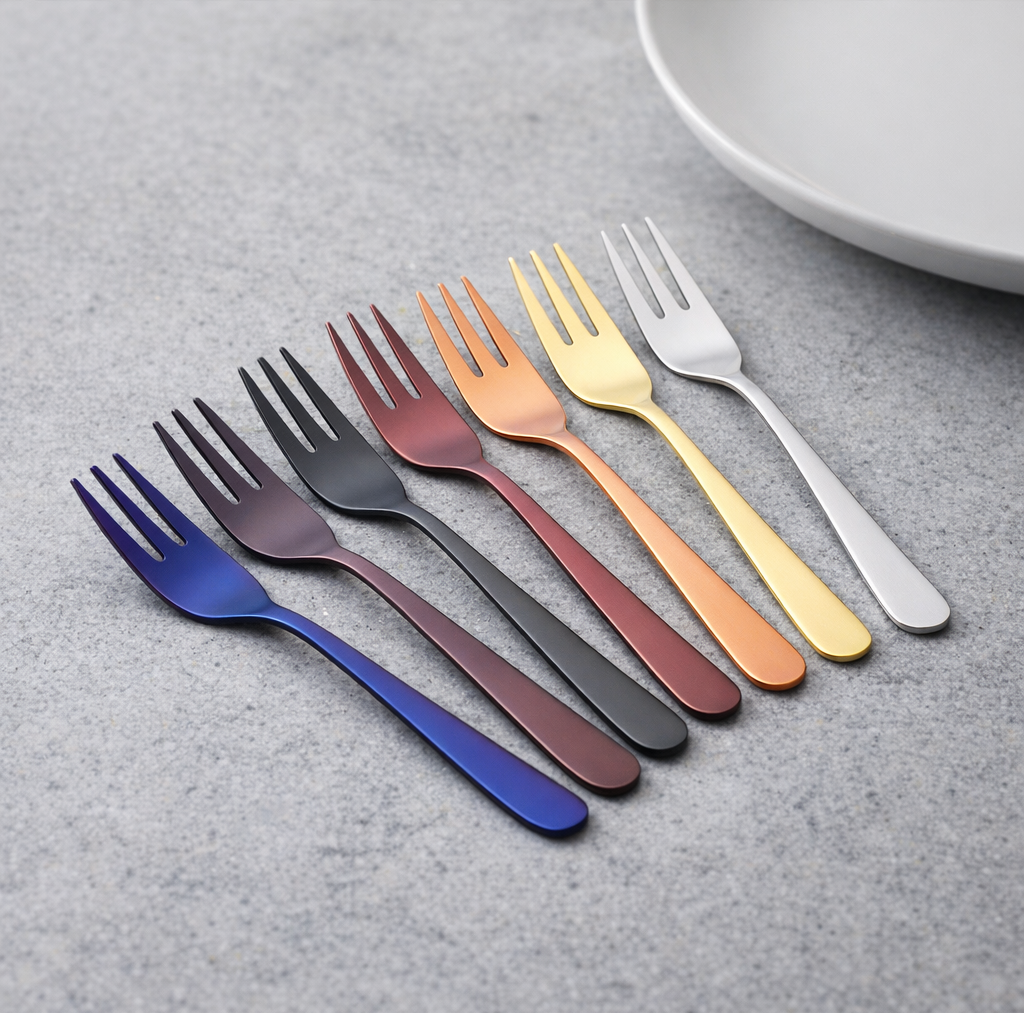 Row of colored salad forks in blue purple gray copper gold and silver finishes laid on a gray textured surface with a partial plate visible in the upper right corner