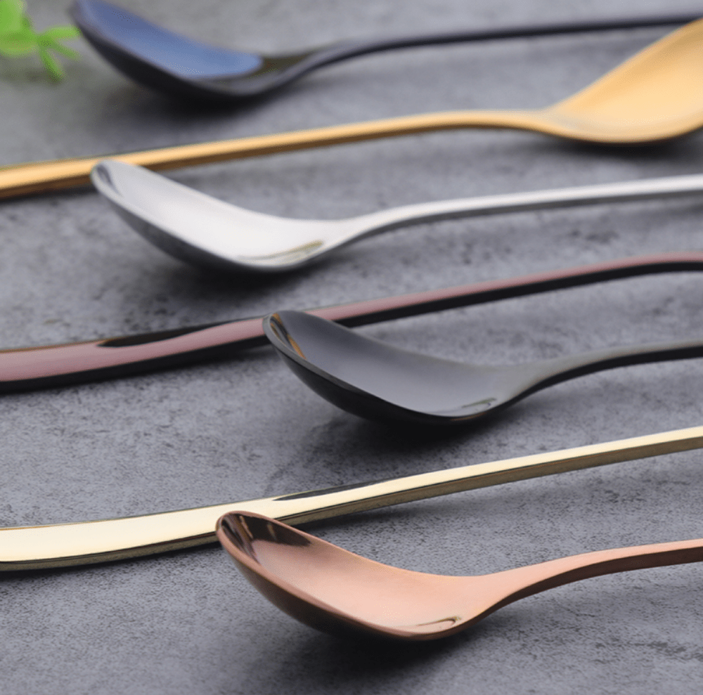 Several dessert spoons with sleek modern handles in blue black silver gold rose gold and copper tones laid on a gray textured surface.
