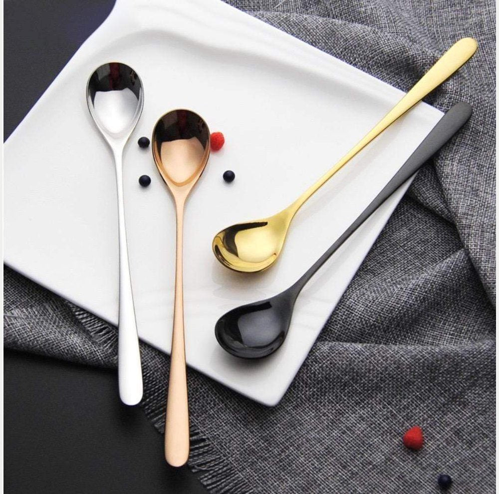 Four dessert spoons in silver rose gold yellow gold and black finishes arranged on a white plate with a gray napkin and small red and dark beads nearby