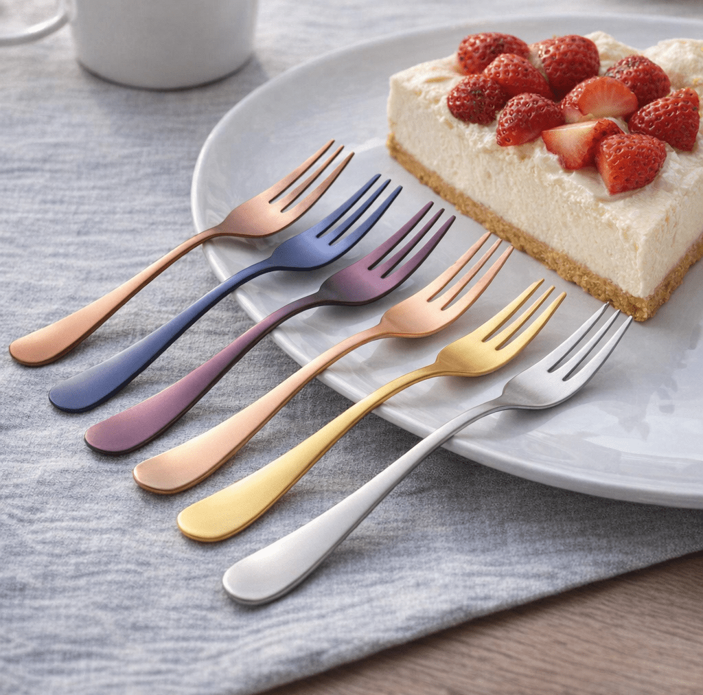 Six brushed colored fruit forks in copper blue purple rose gold and silver rest on a light plate beside a strawberry cake slice