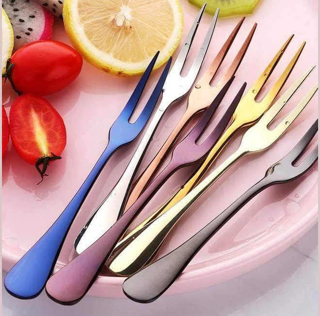 Colorful brushed stainless steel fruit forks arranged on a pink plate with a lemon slice and red tomatoes