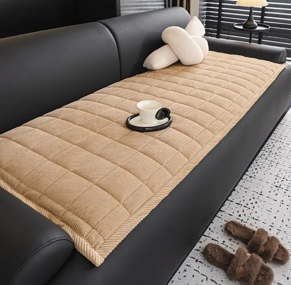 Beige quilted microfiber sofa cover mat with non slip backing on a black leather sofa with a white cup on a saucer a plush pillow a side table lamp and brown fuzzy slippers on a light speckled rug