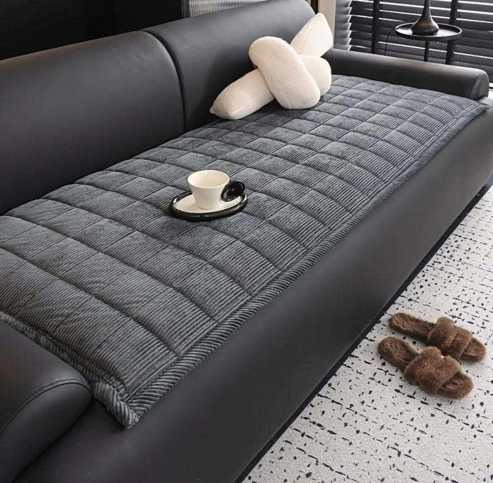 Harborline Quilted Sofa Cover Mat in gray microfiber with non slip backing laid over a black sofa, with a cup on a tray, white pillows, and brown fuzzy slippers on a speckled rug near window blinds