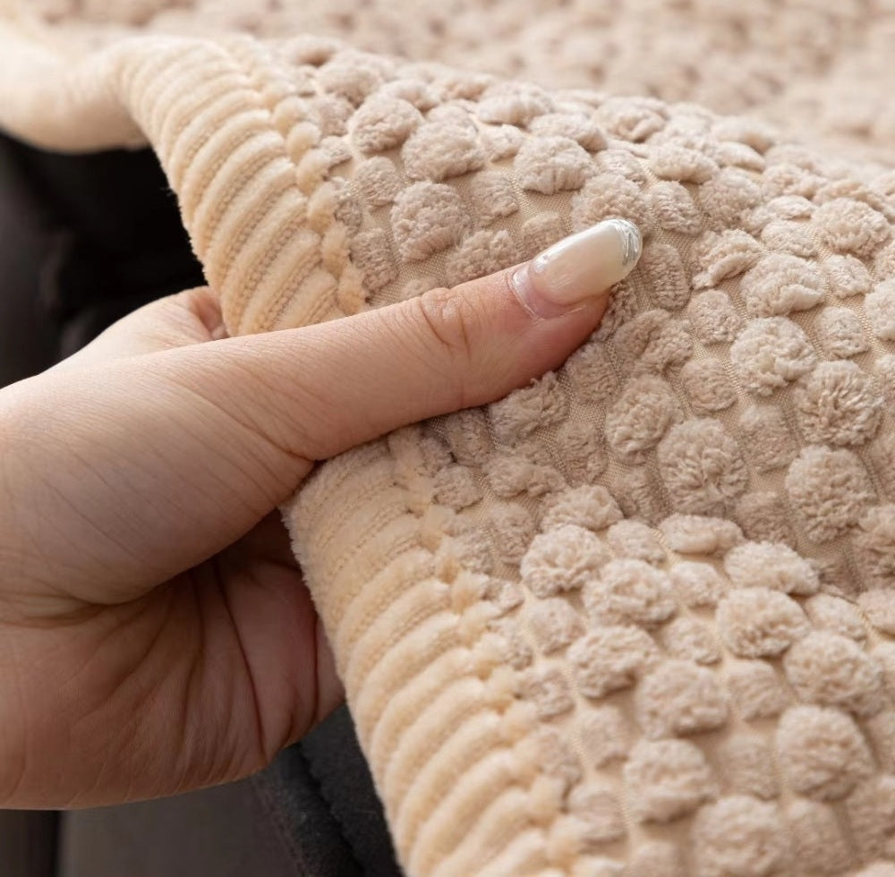 Close up of a beige textured sofa protector slipcover held by a hand, featuring small pom pom tufts and a ribbed edge