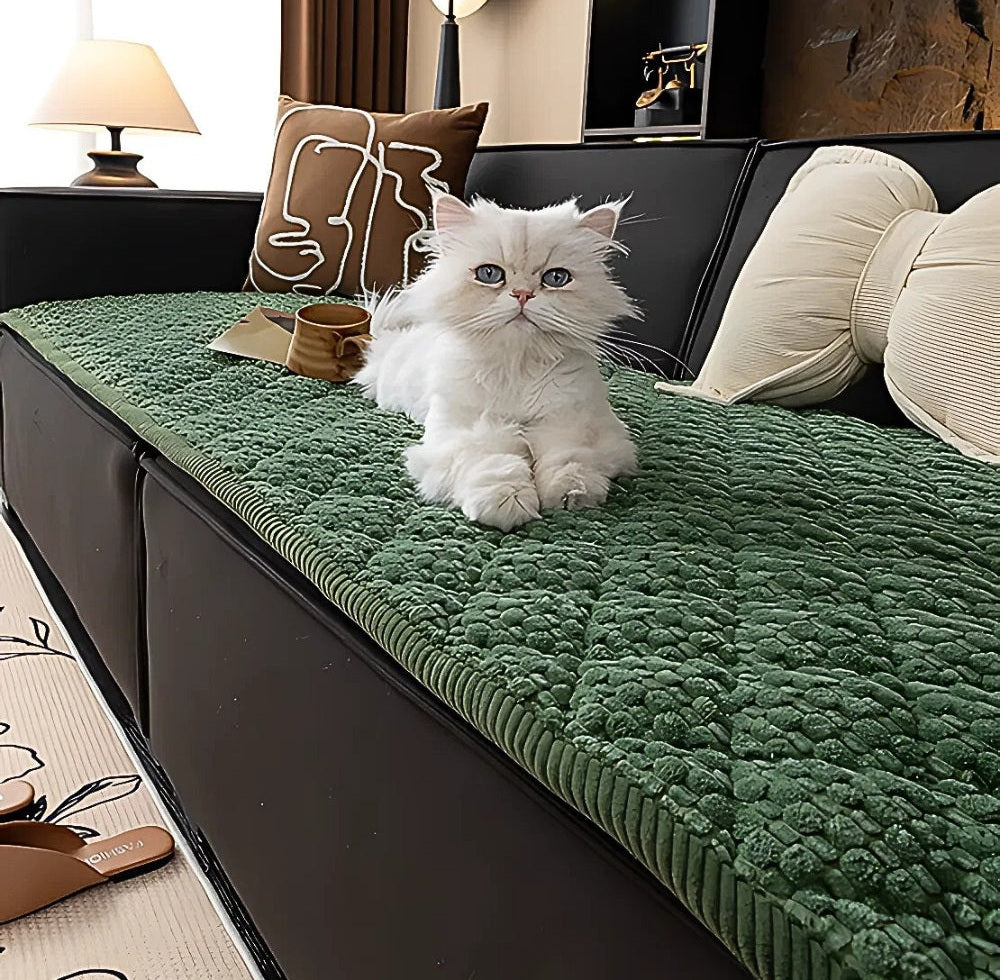 White cat lounging on a green textured quilted sofa protector slipcover over a dark sofa with beige cushions and a lamp in the background