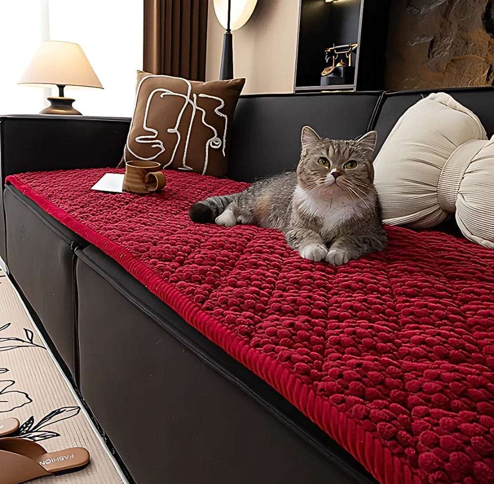 Havenweave sofa protector slipcover in red textured microfiber with non slip backing on a black sofa, a cat lounging on top, brown decorative pillow with white abstract lines and beige cushions, a mug and a note on the cover, and a lamp on the side table in a living room