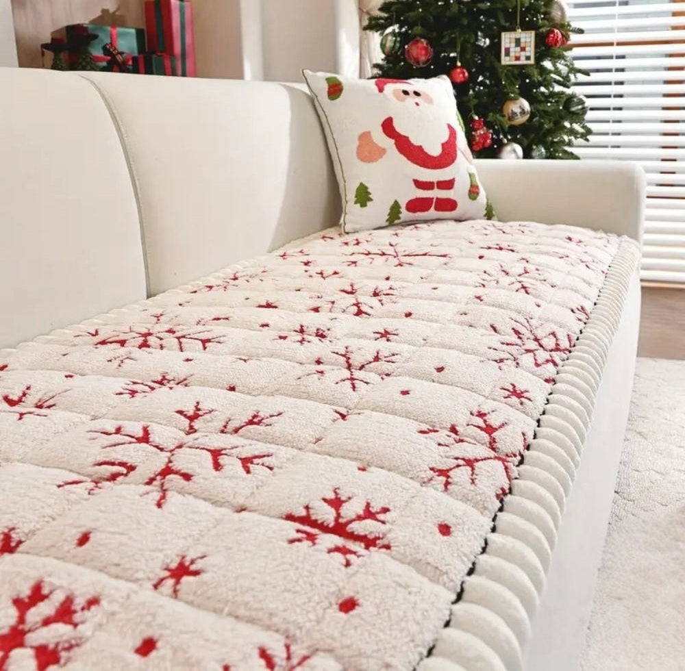Cream sofa with a thick padded cushion style cover showing red snowflake patterns, a Santa Claus pillow, and a Christmas tree with ornaments in the background.