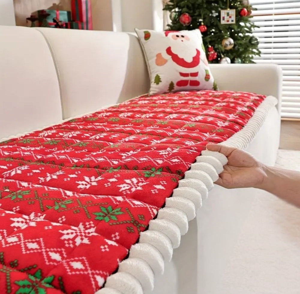 White sofa with a red quilted Christmas sofa cover featuring white snowflake patterns and a white pom pom trim, a Santa pillow and a Christmas tree in the background