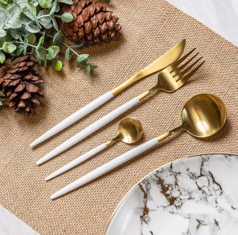 Gold plated cutlery set with white handles including a knife, fork, and two spoons arranged on a burlap placemat with pinecones and greenery and a marble plate edge