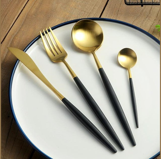 Gold plated cutlery with black handles including a knife, a fork, and two spoons arranged on a white plate with a blue rim on a wooden table