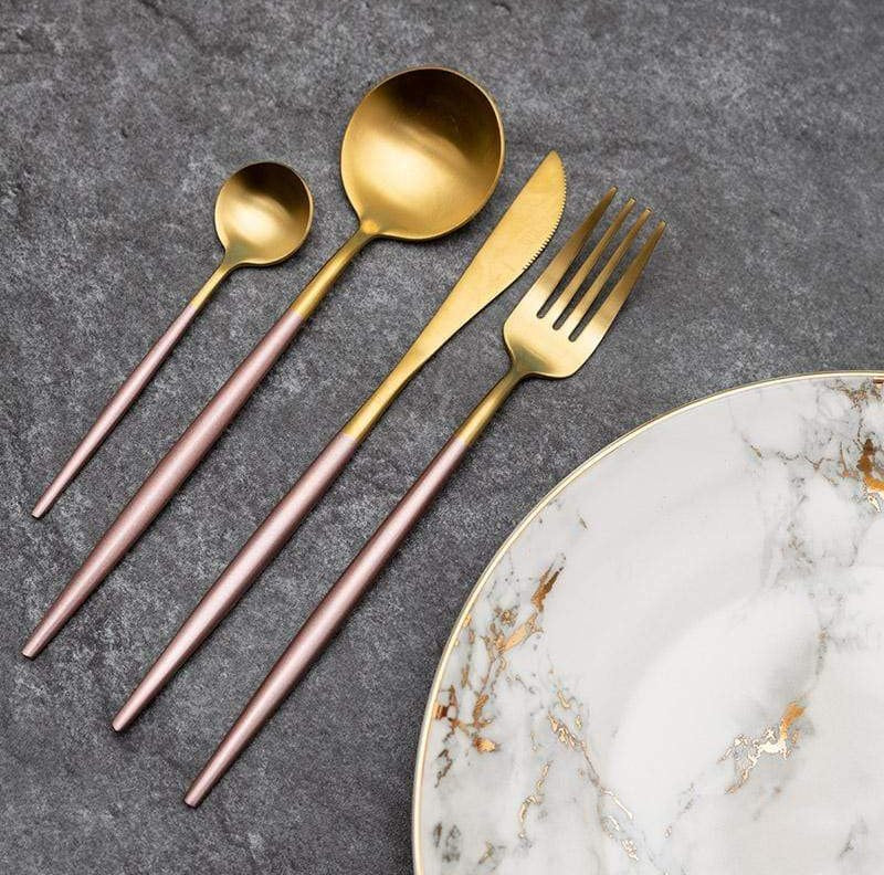 Gold and rose gold plated cutlery set with a small spoon, a large spoon, a serrated knife, and a fork arranged on a dark gray surface beside a marble plate with gold veining.