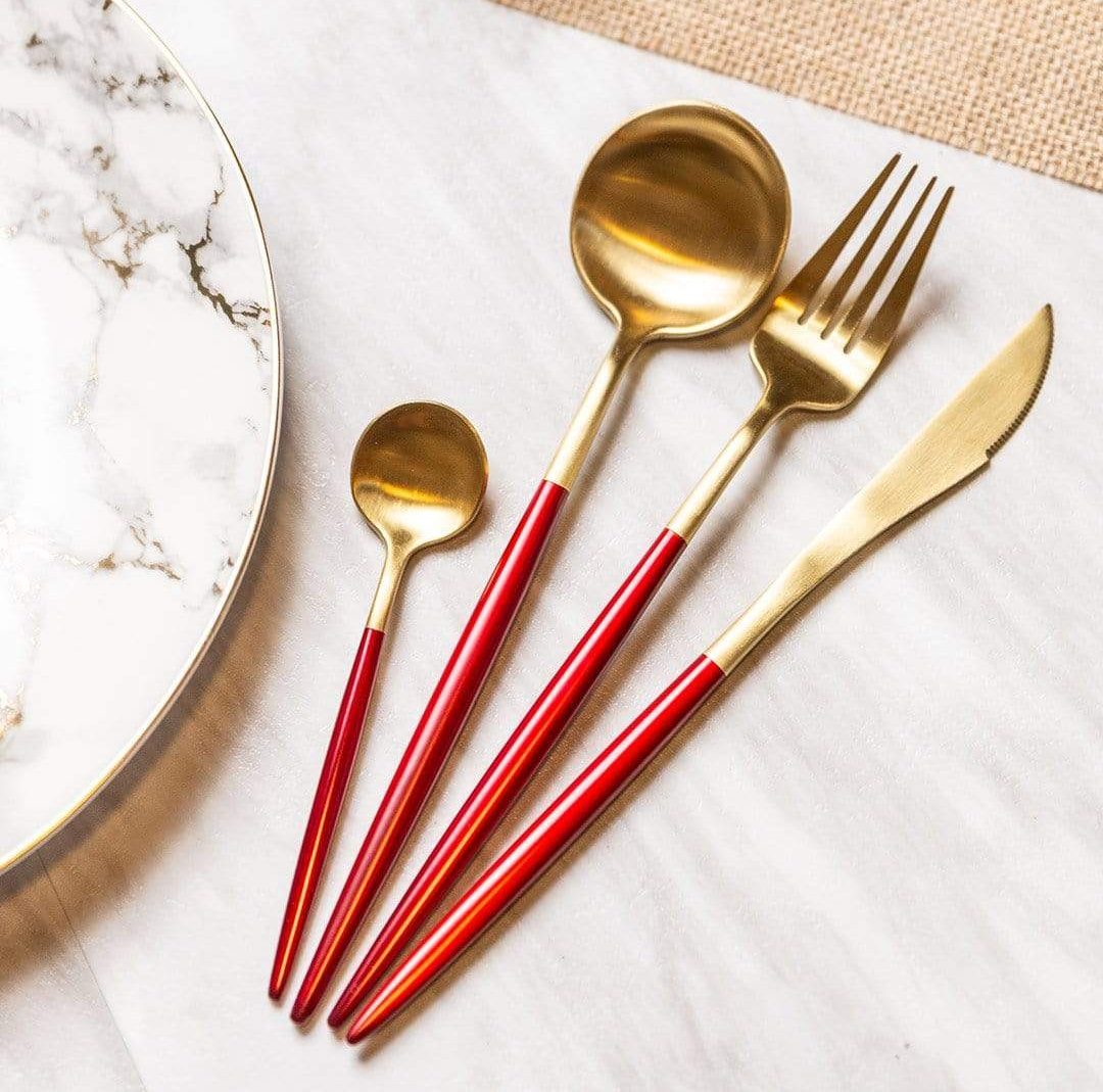Four cutlery pieces with red handles and gold ends including a small spoon, a large spoon, a fork, and a serrated knife on a white marble surface beside a marble plate