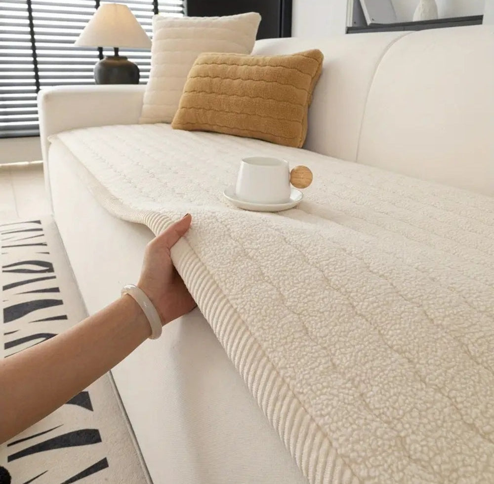 Cream plush sofa cover with a textured surface draped over a white sofa, a hand pulling the edge, beige and white pillows, and a white cup on a saucer with a small wooden spoon beside it.