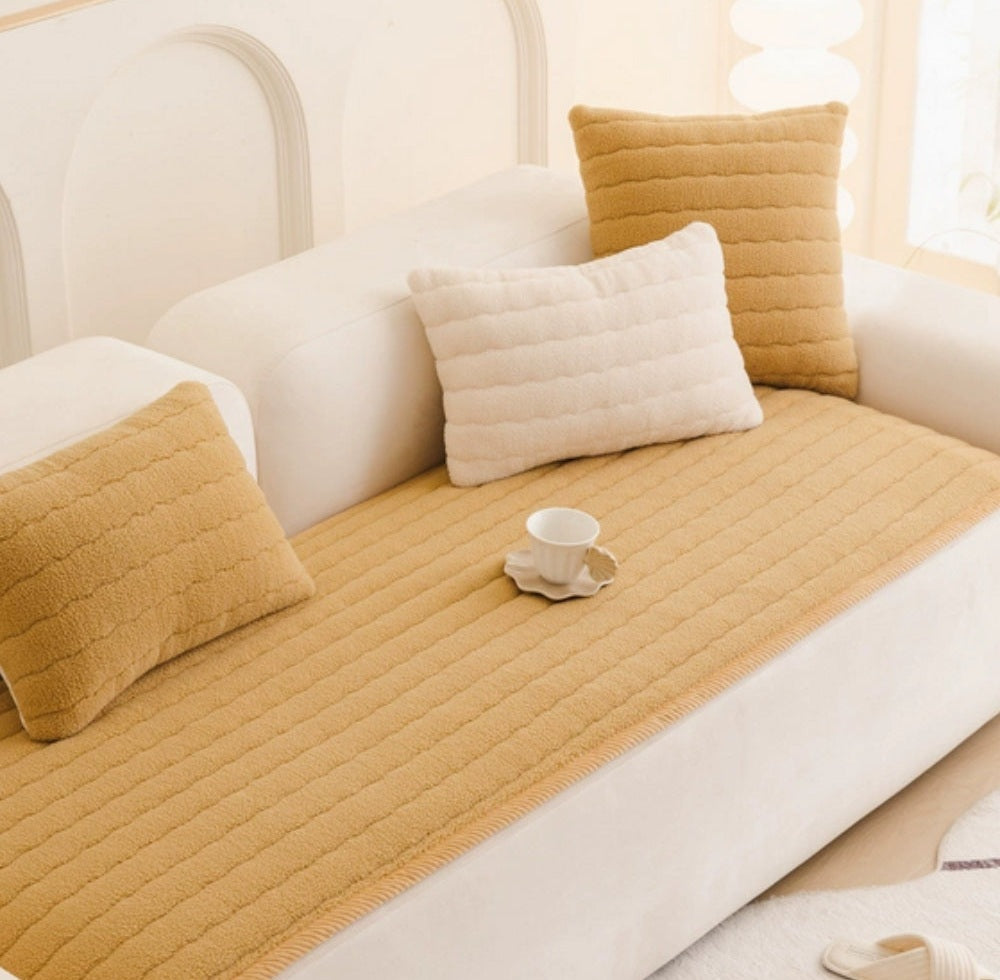 White sofa with a beige plush quilted cover, two tan cushions and a white pillow, and a small cup on a saucer resting on the cover.