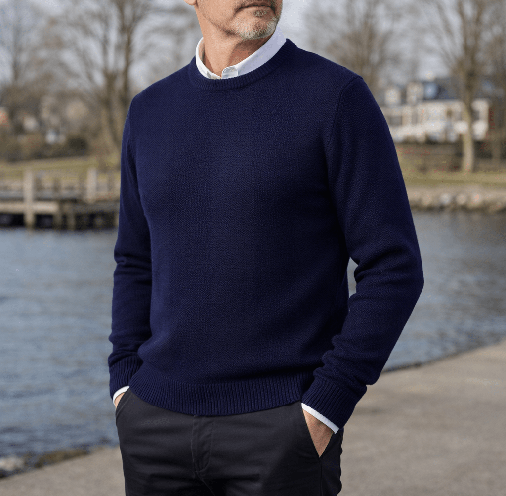 Man wearing a navy blue cashmere pullover sweater with a classic crew neck, white shirt collar visible, standing by a waterfront