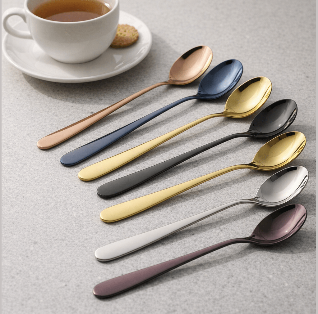 Set of stainless steel spoons with plated finishes in rose gold, blue, gold, black, silver, and purple arranged diagonally on a gray surface beside a white cup and saucer with a cookie