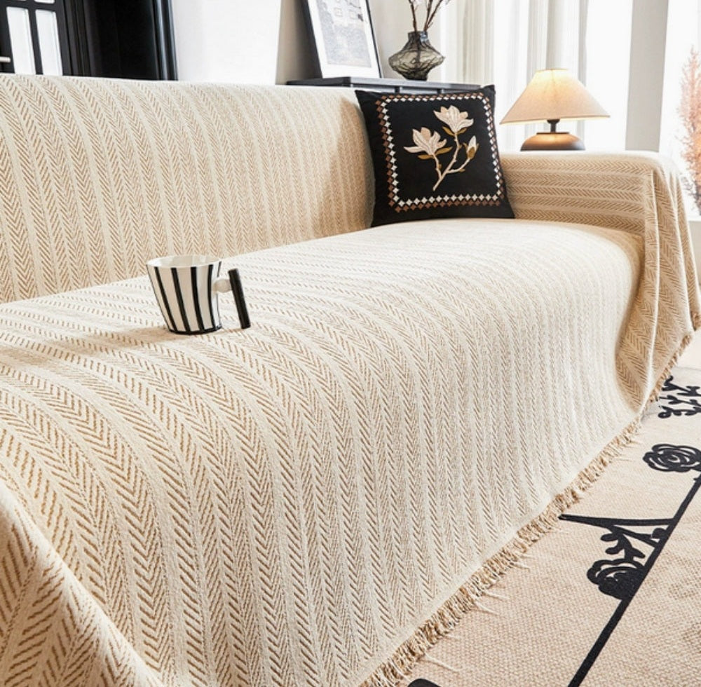 Beige sofa covered with a herringbone chenille throw featuring tassel trim, a black embroidered cushion on the back, and a striped mug resting on the seat.