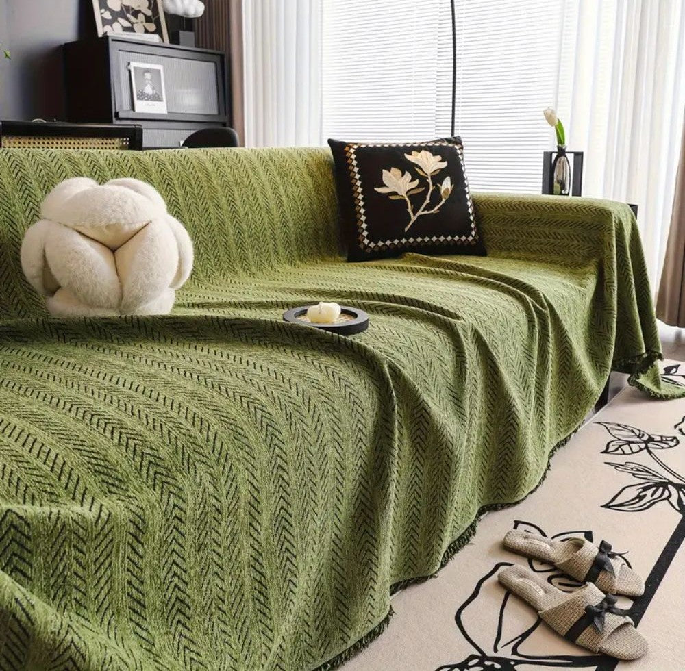 Green herringbone microfiber blend chenille sofa cover with tassel trim draped over a sofa, with a cream knot pillow and a black floral cushion, a candle on a tray, and woven slippers on a light rug.