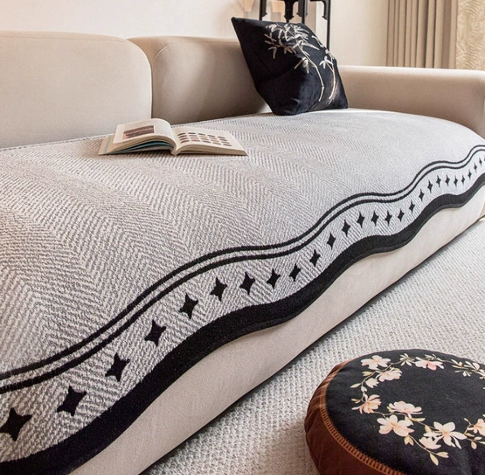 Seabrook sofa cover in light gray microfiber with a black wavy edge border on a beige sofa, an open book resting on the cover and a dark decorative pillow nearby