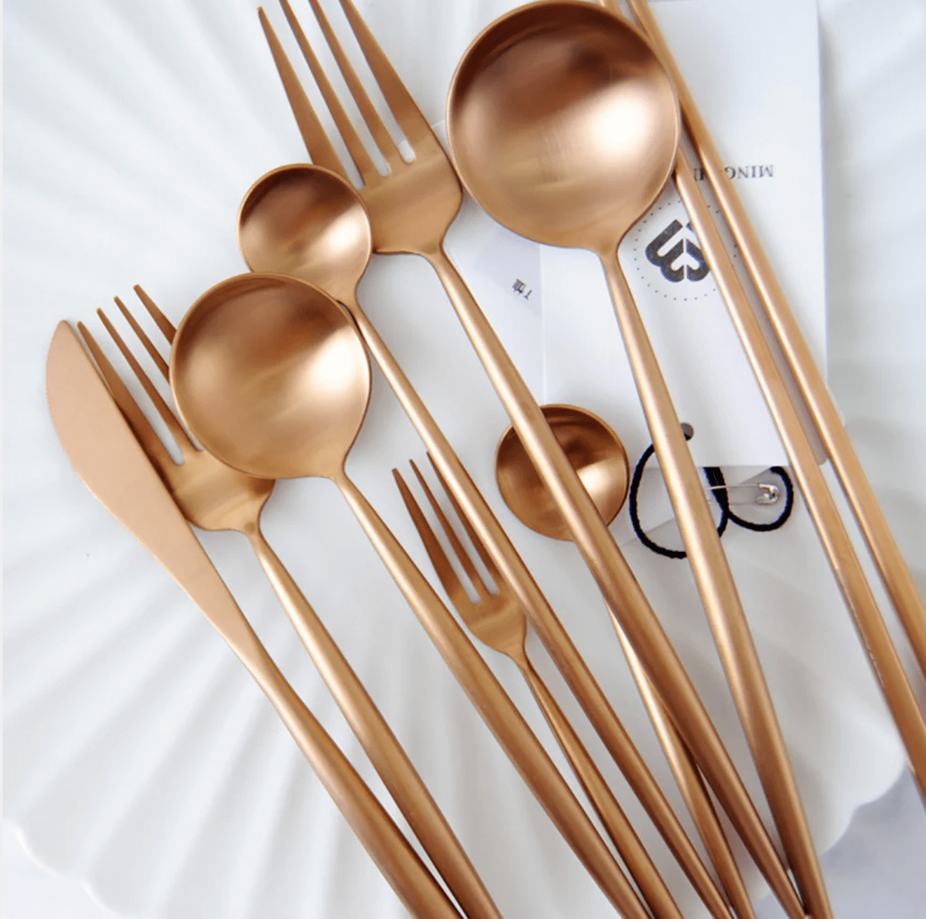 Sevilla Curve stainless steel cutlery set with slim curved design in copper plated finish laid out on a white surface including forks spoons knives and a ladle