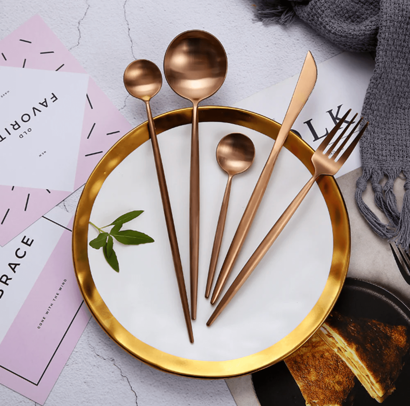 Sevilla Curve copper plated stainless steel cutlery set with slim curved handles displayed on a white plate with a gold rim including a knife a fork and several spoons