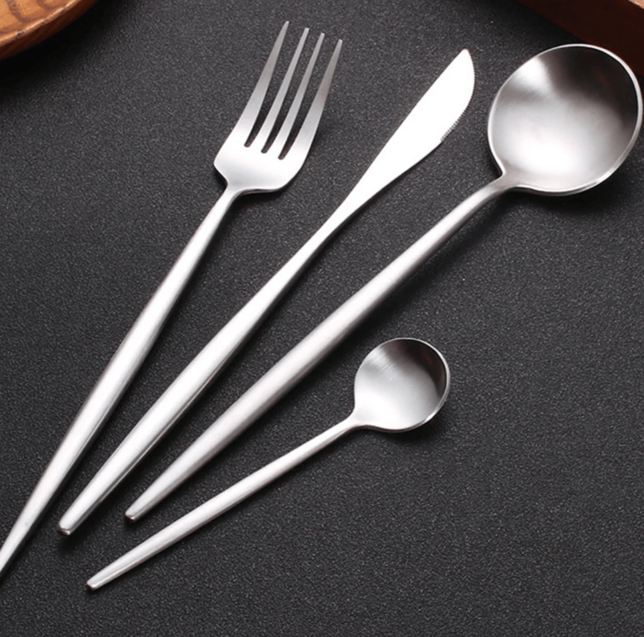 Sevilla Curve stainless steel cutlery set with slim curved handles showing a fork a knife and two spoons on a dark textured surface