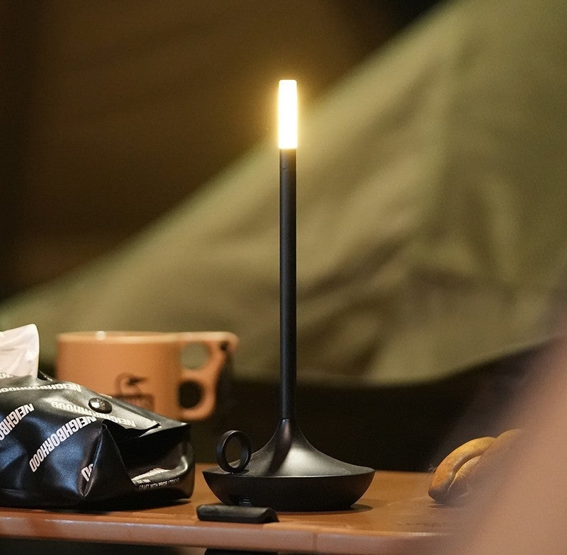 Tall black LED table lamp with a glowing rectangular top on a curved base sits on a wooden table, with a peach colored mug, a black bag with white text, a small remote, and a croissant nearby.