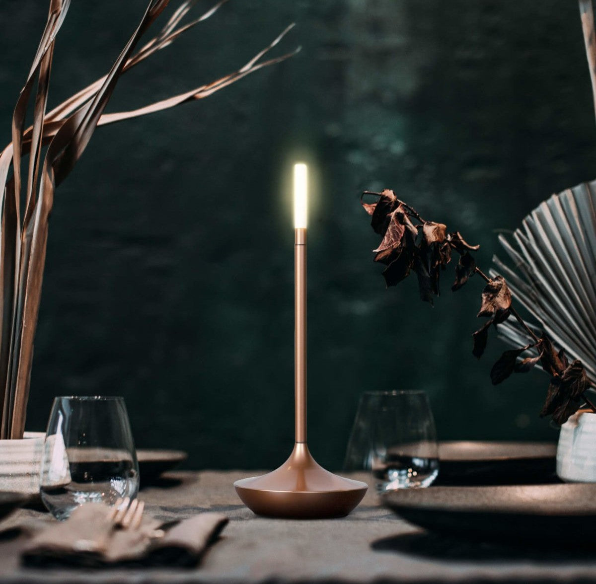 Slender copper table lamp with a bright vertical LED on top resting on a round base on a dining table with glasses and dried leaves