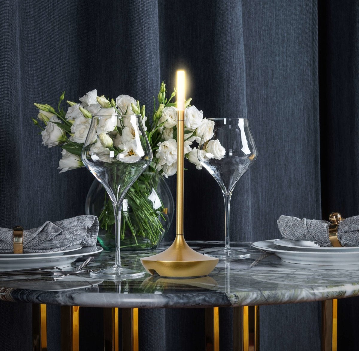 A gold brass plated LED table lamp with a candle like glow sits on a marble dining table beside a vase of white flowers and two wine glasses with gray napkins and gold napkin rings against dark blue curtains