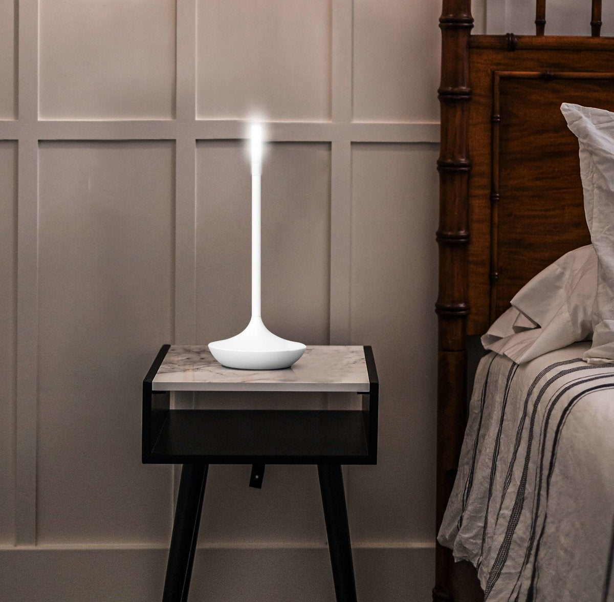 White modern LED table lamp with a tall slim stem and round base on a marble top bedside table beside a bed with a wooden headboard, lit.