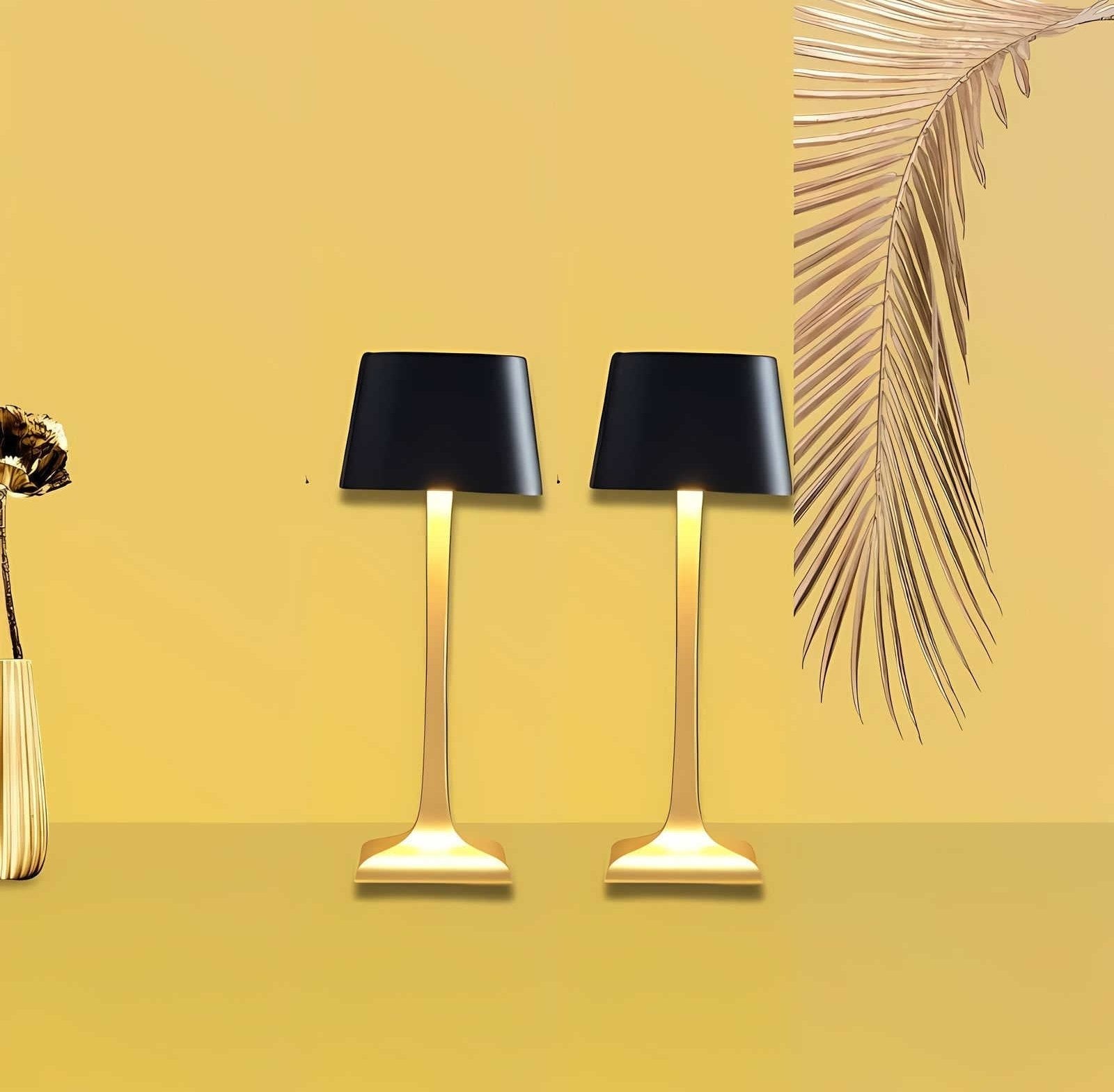 Two cordless table lamps with black shades and gold stems on a yellow background, with a vase on the left and a palm frond on the right