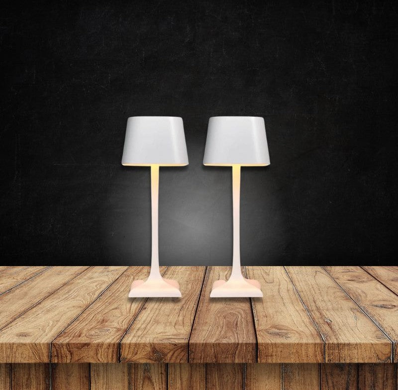 Two white table lamps with drum shades and slender stems on a wooden surface against a dark wall.