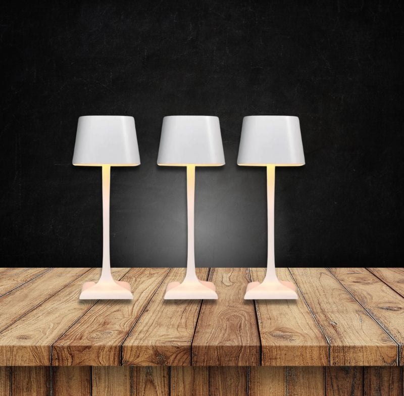 Three white cordless table lamps with slender stems and gray shades arranged on a wooden table against a dark background.