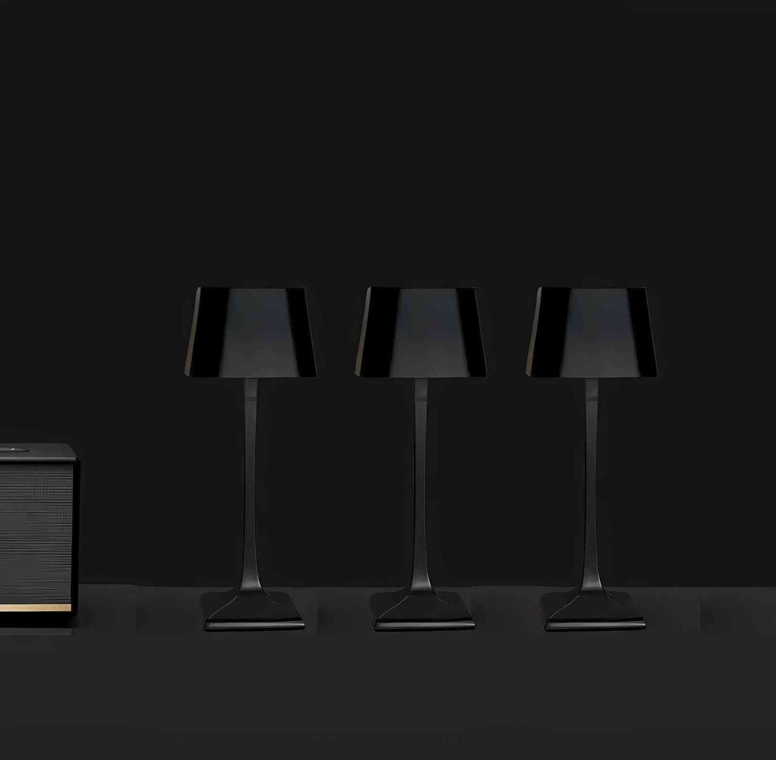 Three black cordless Solara table lamps with square shades and slim stems arranged in a row on a dark surface with a small dark cabinet on the left