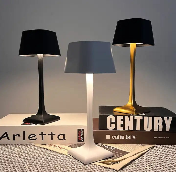 Three cordless table lamps black left white center and gold right sit on a desk beside stacked books and a newspaper