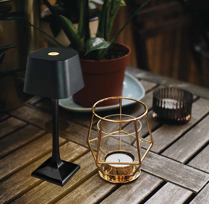 Black cordless table lamp with a round touch button on a square base sits on a wooden outdoor table beside a gold geometric lantern containing a candle and a potted plant in the background