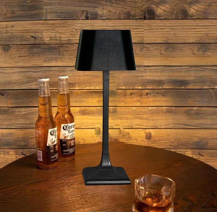 Cordless table lamp with a black shade and slim dark metal stem sits on a round wooden table beside two Corona beer bottles and a glass of whiskey against a wood paneled wall.